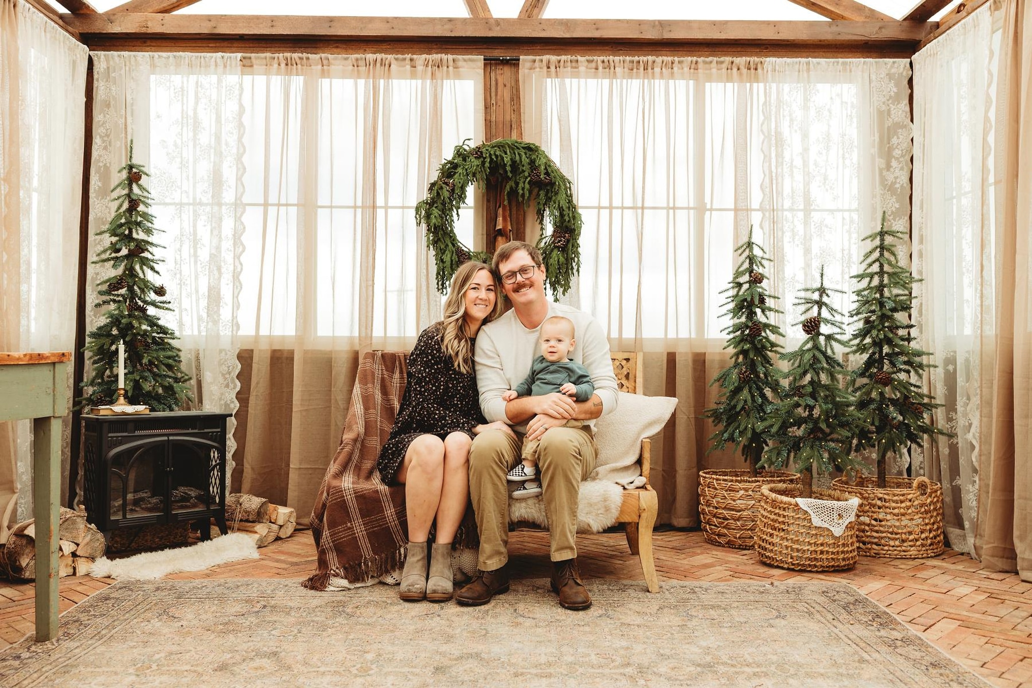 A family seated on a bench beneath an evergreen wreath inside a glass greenhouse strung with lights and lined with little pine trees, photographed by Karismatic Photos